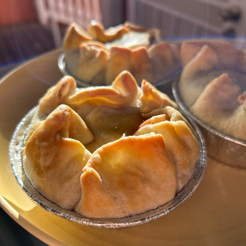 Close-up of several golden-brown HI Pie Bakery + Cafe mini pies in foil cups on a yellow tray.
