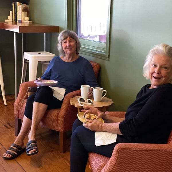 Two smiling women enjoying coffee and pastries in cozy armchairs at HI Pie Bakery + Cafe.