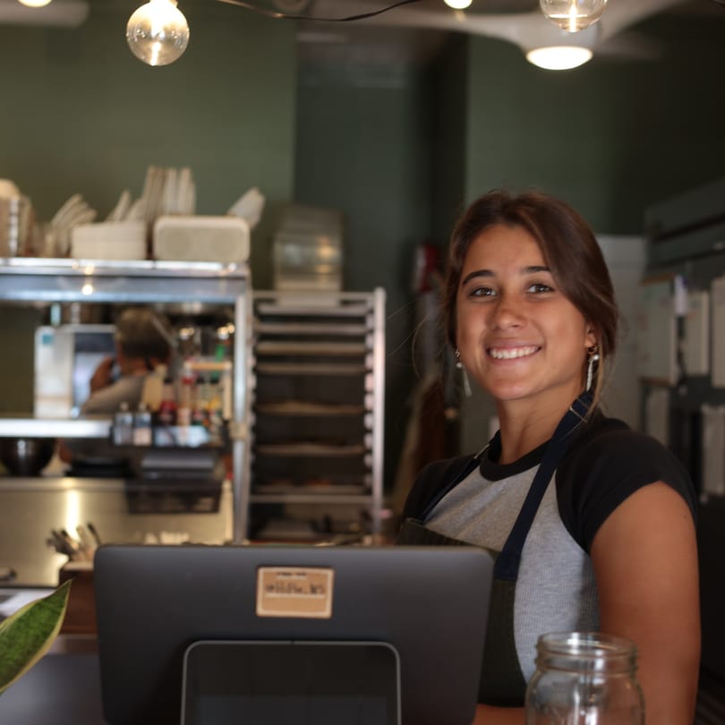 Smiling female employee in an apron stands behind a counter with a register and plant.
