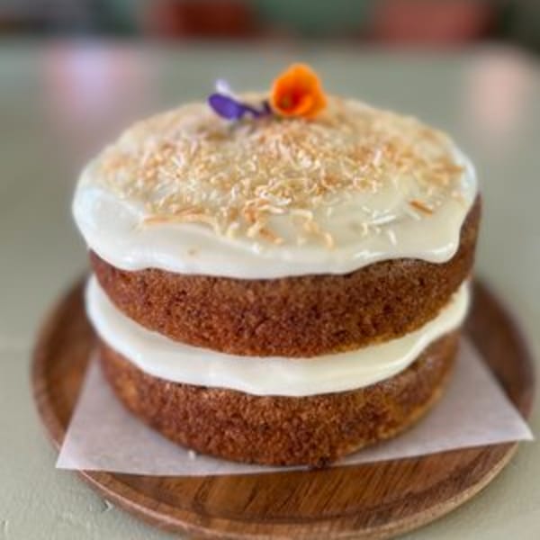 Two-layer carrot cake with white frosting, toasted coconut flakes, and edible flowers on a wooden plate.