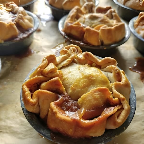 Several golden-brown HI Pie Bakery + Cafe mini pies in dark metal tins, some with filling bubbling over.