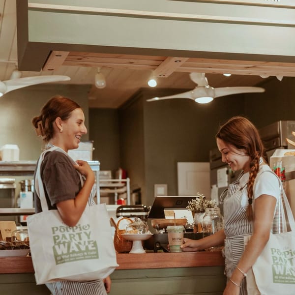 Two women in aprons and carrying "Pot Pie Party Wave" tote bags smile at the HI Pie Bakery + Cafe counter.