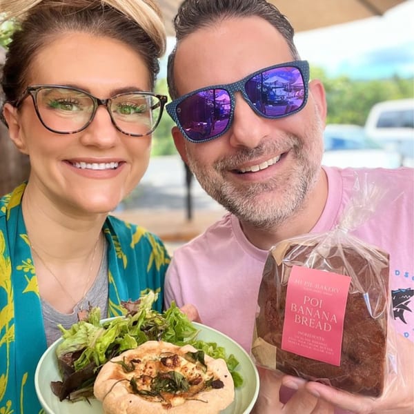 A smiling couple holds a plate with a savory pastry and salad, and a loaf of HI Pie Bakery poi banana bread.