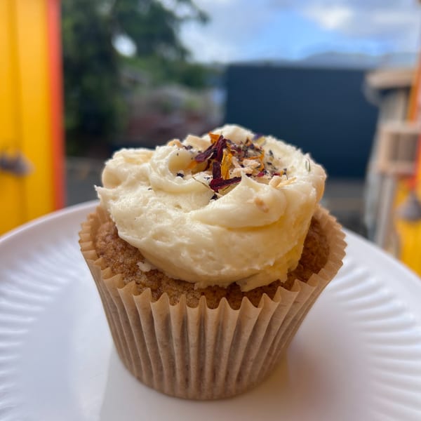 A cupcake with white frosting and colorful sprinkles sits on a white plate outdoors.