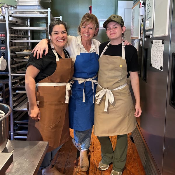 Three smiling HI Pie Bakery + Cafe staff members in aprons pose in a commercial kitchen.