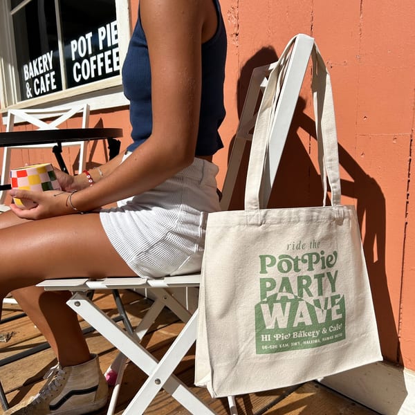 A person sips coffee outside HI Pie Bakery & Cafe, with a branded tote bag on a chair.