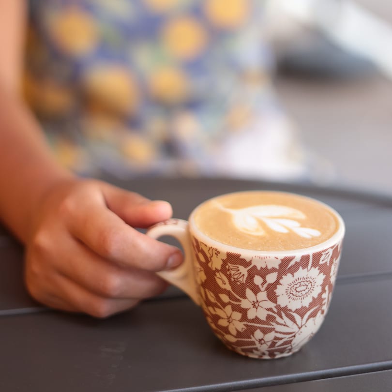 A hand holds a patterned coffee cup with latte art on a dark table.